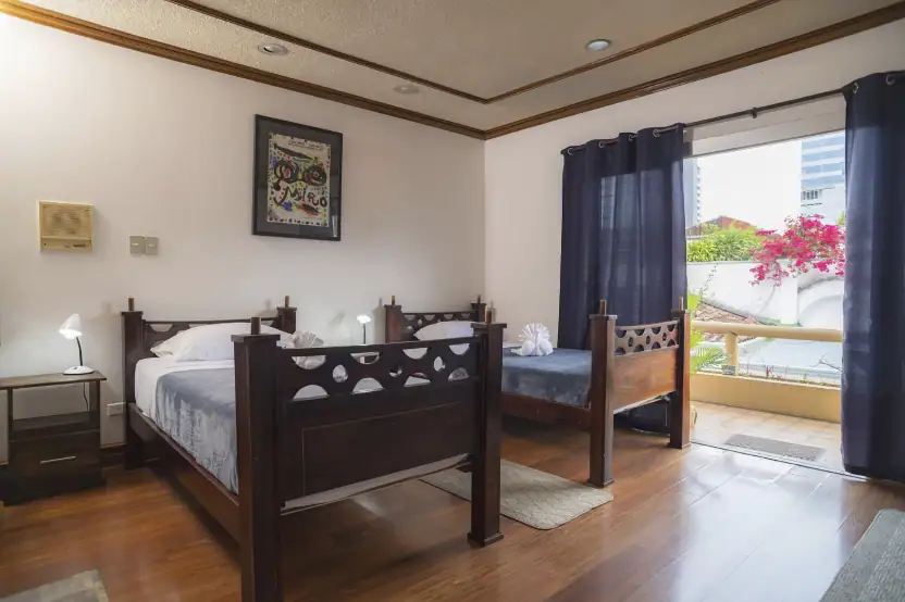 Spacious bedroom with a dark wooden bed, bedside lamps, framed wall art, and blue curtains framing a window that opens to a balcony with greenery and pink flowers.