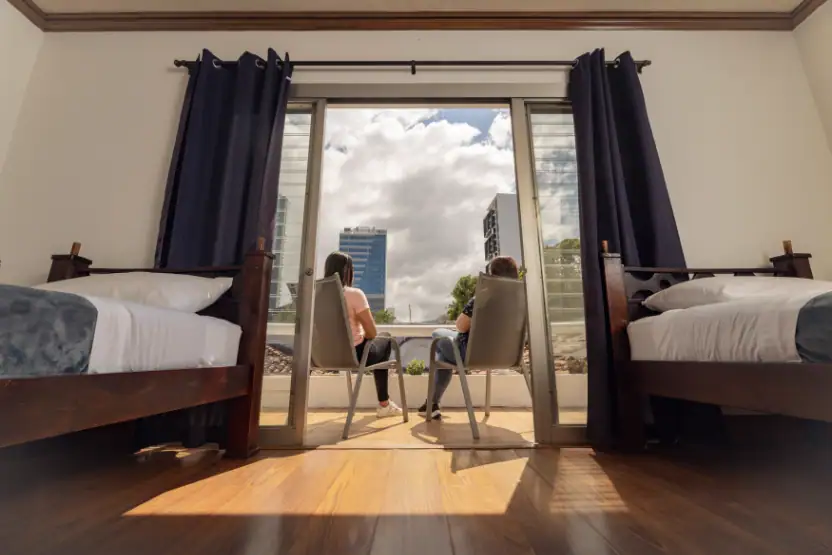 View from inside a room with two beds and dark curtains framing an open sliding glass door onto a balcony. Two people stand on the balcony, seated chairs in view, with tall city buildings and clouds beyond.