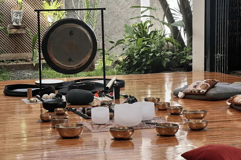 Tea ceremony setup on a wooden table outdoors with multiple small bronze cups, a white serving pitcher, black tea tools, and incense items, with a small hanging calligraphy sign in the background among plants and a stone wall.